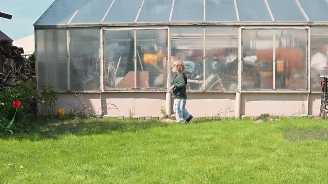 A young child, approximately four years old with light curly hair, runs across a grassy area while playing with a ball
