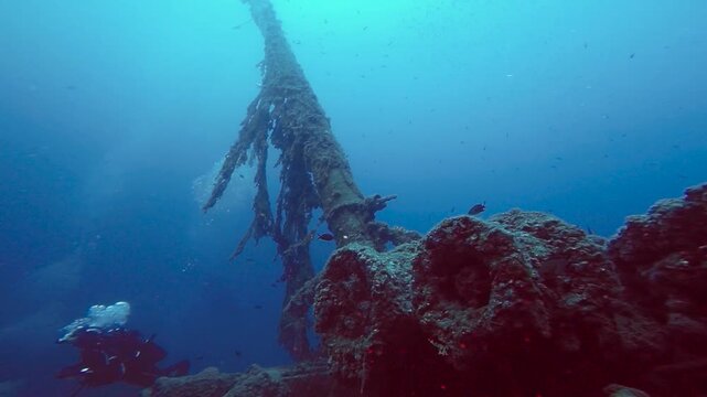 Cinematic Ascent Through Sunken Ship Frames to Diver Hovering Over Deck. A slow motion underwater shot starting with a low angle of a massive, 25-meter rusted ship mast surrounded by deep blue water. 