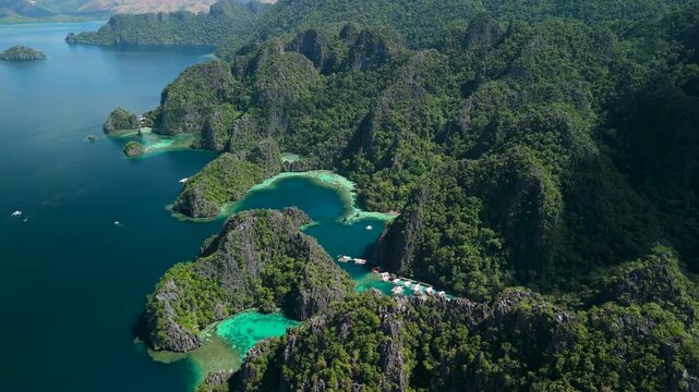 Aerial flypass above limestone karst mountain tops revealing hidden entrance to Kayangan Lake with many parked outrigger tour boats in Coron Palawan Philippines