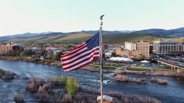 Large American flag on pole overlooking Missoula downtown, Clark Fork River with rapids, road bridge, riverside park and snow-capped mountains under clear blue sky. Montana, USA. Aerial orbit wide.