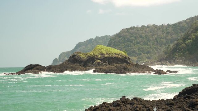 Waves crash on rocky shoreline with lush island and forested hills in background