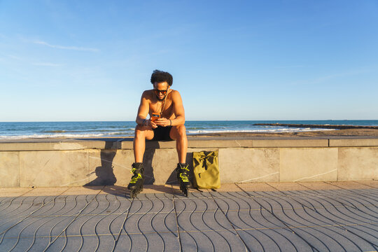 Young man sitting on a wall scrolling phone and wearing rollerblades at the beach
