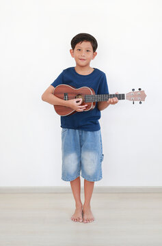 Portrait of happy young Asian boy playing the ukulele, learning a new hobby at home against white wall.
