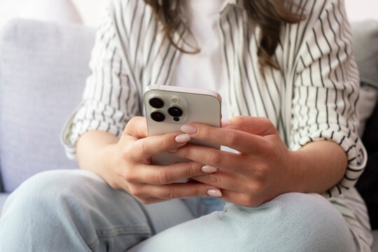 A relaxed woman sitting on a couch holding a smartphone in both hands, scrolling and texting while wearing a striped shirt and jeans, focused on social media, communication and digital lifestyle.