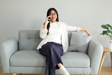 Obraz premium A young woman is sitting on the sofa at home, making a call using her smartphone, chatting or communicating with someone.