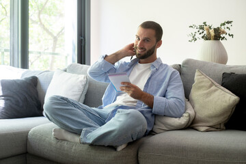 Obraz premium Smiling man sitting cross-legged on a gray couch at home, talking on the phone and taking notes in a spiral notebook while relaxed in casual clothes in a bright living room