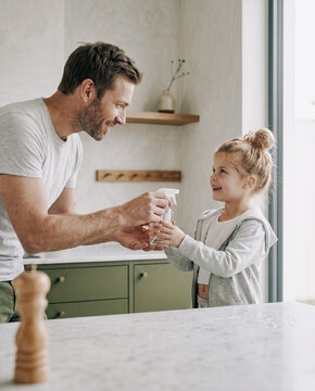 An intimate waist-up shot of a father handing a minimalist glass spray dispenser to his smiling daughter, captured against dark olive cabinets in bright morning rim light.
