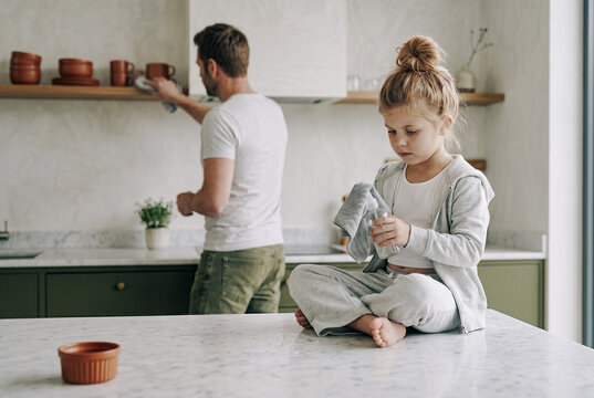 A young daughter sits cross-legged on a veined marble island cleaning an object, while her father wipes an open oak shelf holding terracotta dishware in the soft background.
