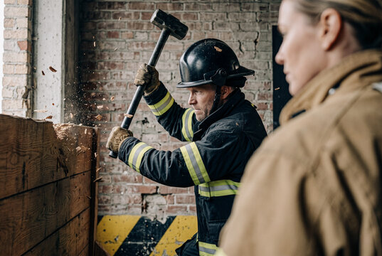 Over-the-shoulder horizontal action shot. Firefighter in de-branded gear is in mid-swing with a massive sledgehammer overhead, muscles strained. Wood fragments and high-energy impact focus.
