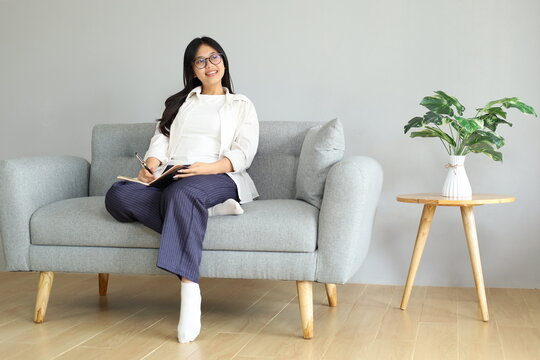 A young woman relaxes sitting on the sofa at home, occasionally looking up, remembering something and writing in a notebook, spending her free time in the living room.