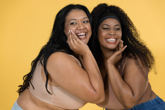 Diverse mid-adult female friends posing close smiling in studio tan bralettes denim yellow backdrop
