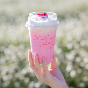 A hand holding a clear plastic cup of pastel pink iced drink topped with thick white milk foam and a tiny pink flower against a white flower field.