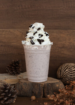 Cookies and cream milkshake smoothie in a plastic takeaway cup topped with whipped cream and crushed chocolate cookies on a rustic wood slice.