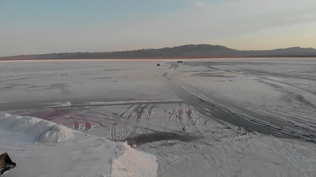 Parallel tracks mark the white salt flat with machinery working in distance under bright sky creating patterned industrial harvesting landscape ideal for mineral production and desert resource stock