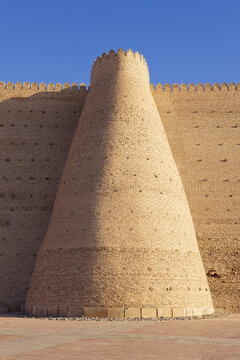 Conical tower and crenellated wall of the Ark Fortress in Bukhara, Uzbekistan. Massive mudbrick fortification under clear blue sky, ideal for travel and heritage publications.