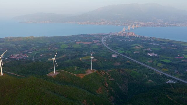 Picturesque aerial view of a wind farm on a lush green mountain with the iconic rio-antirrio bridge spanning the gulf of corinth in the background, showcasing renewable energy production in greece