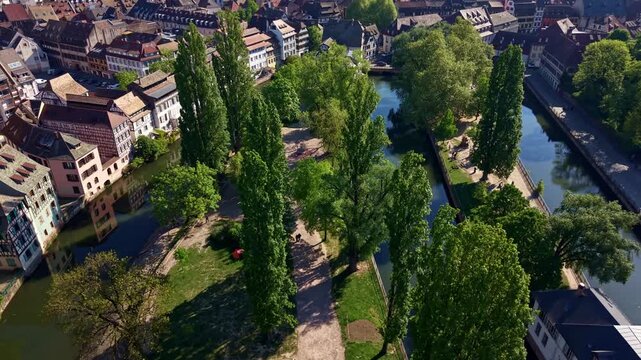 Drone backward fly over lush green trees lining near urban pathways along the ill river canals in Strasbourg's Square Louise-Weiss area, Petite France, France