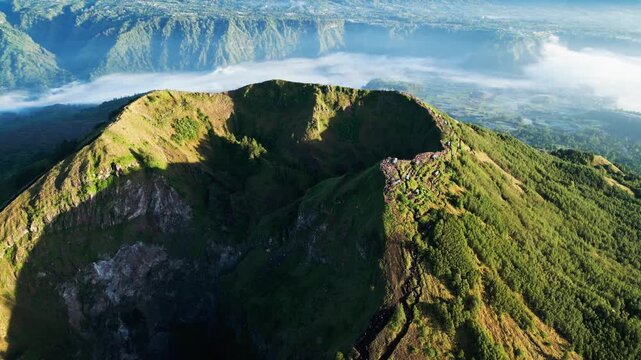 Mount Batur crater rim glowing in golden morning sunlight above mist covered caldera valley and rugged volcanic landscape orbit drone footage.