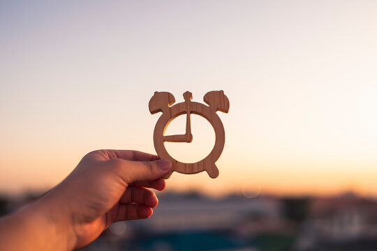 Hand holding a wooden alarm clock shape against a blurred warm sunrise sky. Concept of time management, morning routine, deadline, future planning, and starting a new business day.