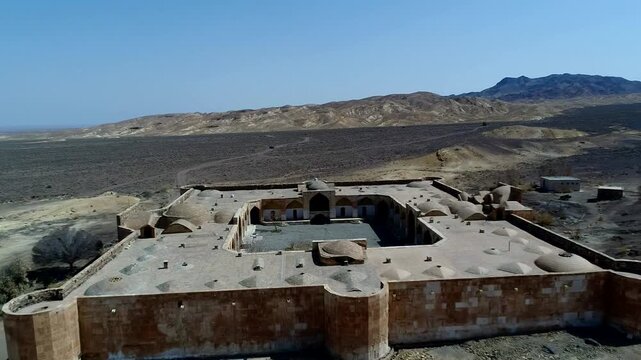 Interior courtyard of stone caravanserai ruins reveals arched walls and open space under bright blue sky in arid desert setting forming atmospheric historical heritage scene perfect for archaeology