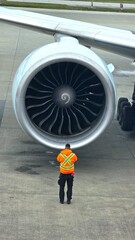 High- angle view of an airport ground crew worker in a high-visibility safety jacket standing in front of a massive commercial airplane jet engine. © Chaetan