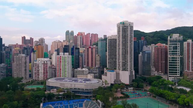 Aerial parallax above Victoria Park swimming pool tennis stadium and colorful basketball courts toward dense Hong Kong residential apartments and skyscrapers against green mountain backdrop