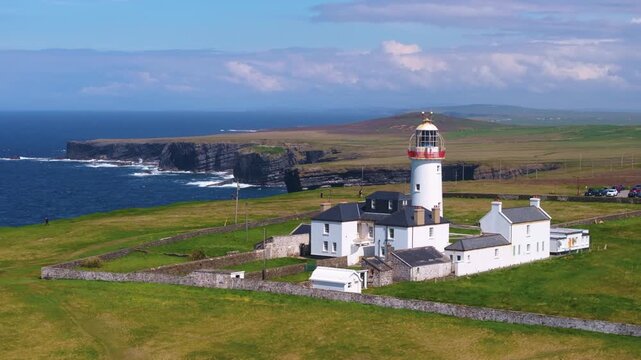 The iconic lighthouse at Loop Head stands with surrounding buildings against a backdrop of ocean and cliffs. Ideal for travel content and heritage documentaries.