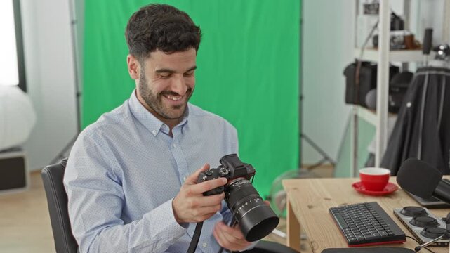 Man holding camera with bare hands checking lens and controls beside desk, keyboard and green screen in studio; satisfaction.