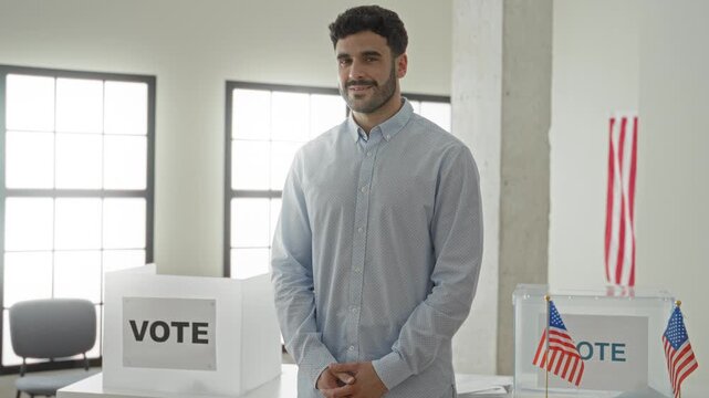 Man smiling with hands clasped by a ballot box and vote sign, standing at a polling table in building; civic duty.