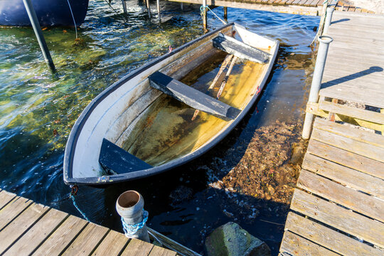 Weathered rowboat filled with shallow water is tied beside wooden docks in a small marina, showing ropes, oars, clear lake water, and rustic boating detail for fishing and travel themes