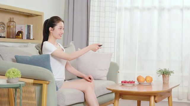 Smiling girl relaxing, using handheld electric mini fan while watching tv in comfortable couch sofa, switching channels using remote controller, choosing programs or shows, enjoying series on weekend