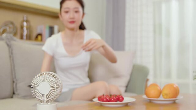 Woman relaxing, using handheld electric mini fan while watching tv in comfortable couch sofa, switching channels using remote controller, choosing programs or shows, enjoying series on weekend home
