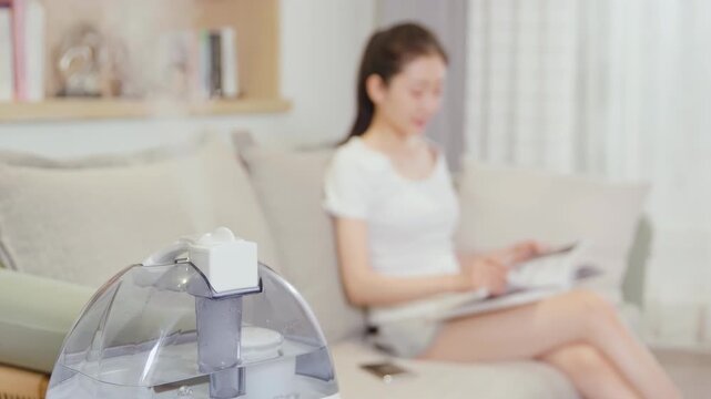 Focused woman sitting on the couch sofa reading a book while, with purifier in room, in smart house. Air cleaning and humidifying device, essential oil diffuser comfortable in living conditions room