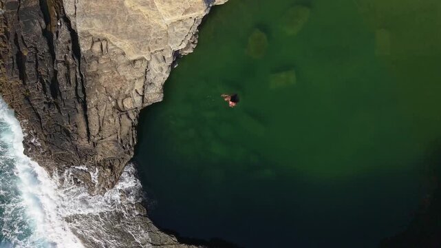 A vertical drone ascent starting with a top-down view of a woman swimming in the emerald waters of the Giola natural pool, rising to reveal the rugged Thasian coastline and Aegean horizon.