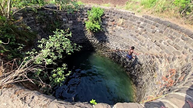 An unrecognized swimmer leaps into a deep stone well in slow motion. They make a huge splash, swim through the dark water, and slowly scramble up the circular, rugged masonry walls.