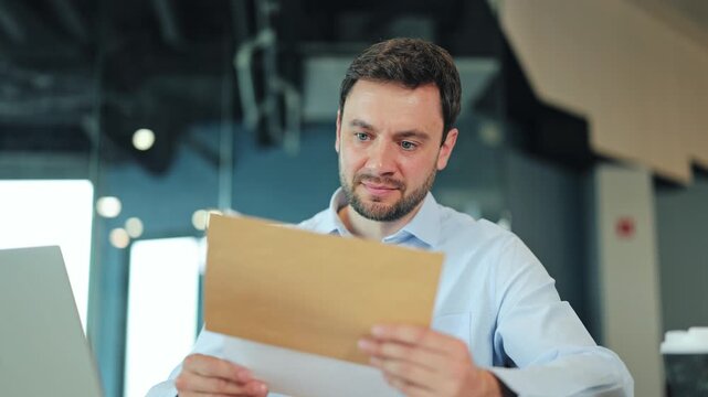 Close-up of smiling businessman or IT specialist checking approved document or offer from envelope. Satisfied corporate worker, manager or freelance designer celebrating success at office desk.
