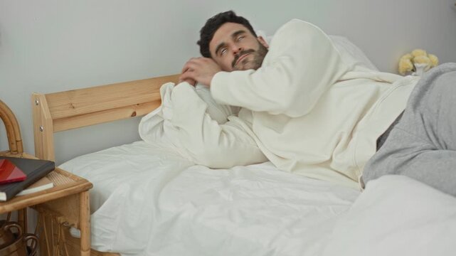 Man hugging pillow and lying on his side on a bed, eyes closed, wearing sweatshirt and sweatpants beside a wooden headboard in a bedroom building; calm rest.