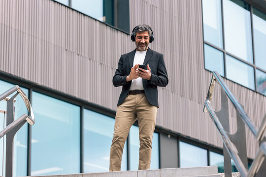 Happy businessman using smartphone while listening to music with headphones in front of the office.
