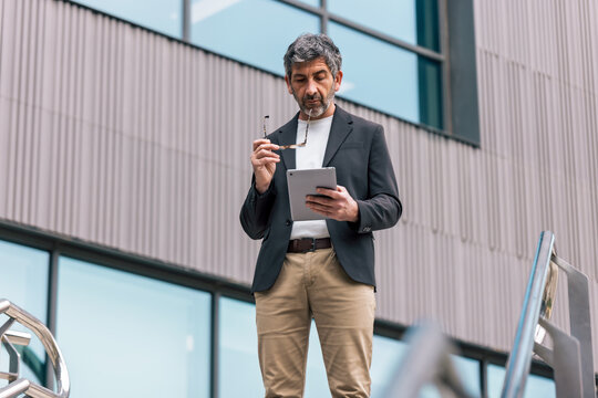 Mature elegant businessman working with digital tablet while staying in front of office