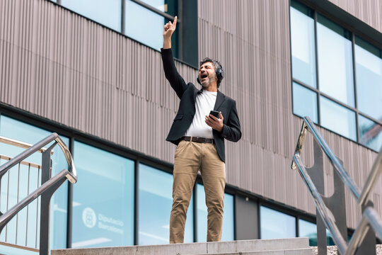 Funny businessman dancing and listening to music with headphones in front of the office.
