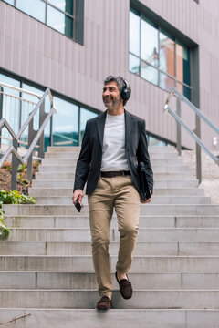 Smiling businessman listening to music with headphones while walking down the stairs in front of the office