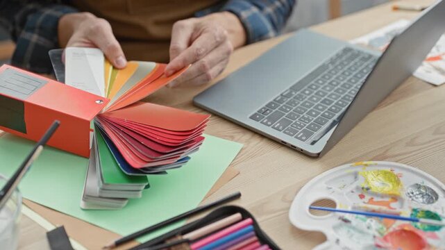 Man painter flipping color swatches with hands beside laptop and paint palette in a studio, wearing apron and checking color samples; creative focus.
