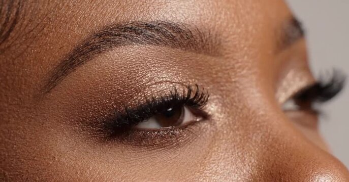 Extreme close-up of eyes of a Black woman with gold shimmer eyeshadow and long eyelashes while blinking.