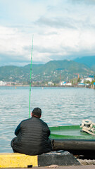 Obraz premium A man sits on a harbor pier fishing in calm waters with scenic mountains behind.