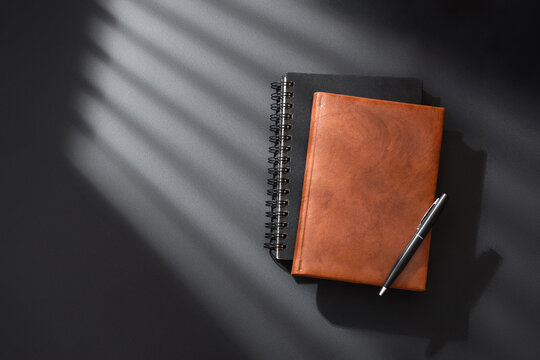 Brown leather and black spiral notebooks with elegant pen on dark surface with striped sunlight shadows from the window. Dramatic workspace flat lay.