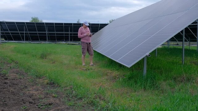 Farmer checking solar panels performance while working in the garden A man with a hoe stands near home solar panels, calculating energy efficiency on a smartphone and taking notes in a notebook. 