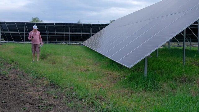 Farmer checking solar panels performance while working in the garden A man with a hoe stands near home solar panels, calculating energy efficiency on a smartphone and taking notes in a notebook. 
