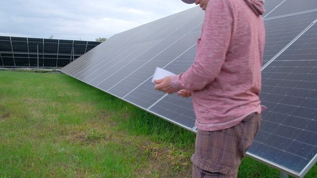 Farmer checking solar panels performance while working in the garden A man with a hoe stands near home solar panels, calculating energy efficiency on a smartphone and taking notes in a notebook. 