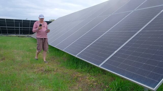 Farmer checking solar panels performance while working in the garden A man with a hoe stands near home solar panels, calculating energy efficiency on a smartphone and taking notes in a notebook. 