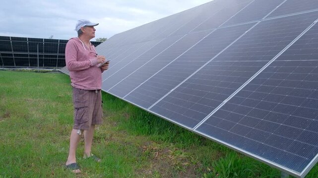 Farmer checking solar panels performance while working in the garden A man with a hoe stands near home solar panels, calculating energy efficiency on a smartphone and taking notes in a notebook. 